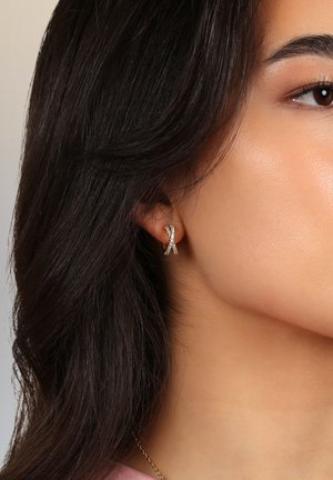 Guldörhänge med vridet ribbdesign, dekorerad med små klara ädelstenar, visad på en models öra mot en neutral bakgrund.