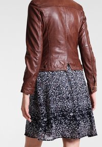 Frau in einer eng anliegenden braunen Lederjacke über einem schwarz-weißen floral gemusterten Rock in knie-langer Länge, von hinten gezeigt.