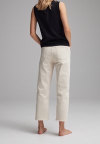Jeans beige a vita alta con orlo grezzo e tasche posteriori, abbinati a un top senza maniche nero. Il tessuto sembra morbido e informale.