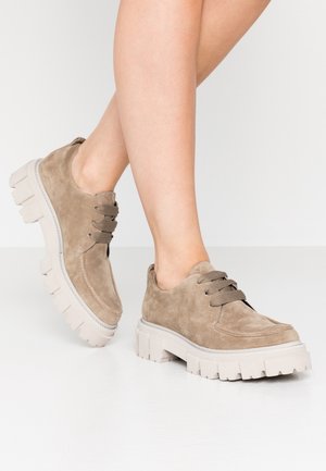Benen met beige suède veterschoenen met dikke, witte zolen, staand op een witte vloer.