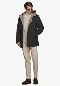 Giacca isolante nera con interno beige, dotata di cappuccio, chiusura a zip e due tasche laterali. Indossata sopra un dolcevita beige e pantaloni chiari.