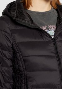 Doudoune noire avec capuche, comportant des sections matelassées et une fermeture éclair à l'avant. Portée sur un t-shirt graphique à encolure ronde.