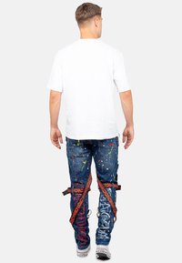 Jeans en denim bleu avec un design de taches de peinture colorées, des bretelles ajustables et un texte graphique contrasté. Associés à un t-shirt blanc uni.
