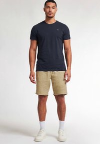 Marineblå bomulds-T-shirt med rund hals, kombineret med lyse beige shorts. Modellen bærer hvide sokker og cremefarvede sneakers og står foran en ensfarvet baggrund.