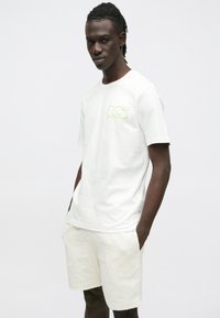 Hvit bomulls-T-skjorte med korte ermer og en neongrønn logo på brystet, kombinert med kremfargede, teksturerte shorts med et geometrisk mønster.