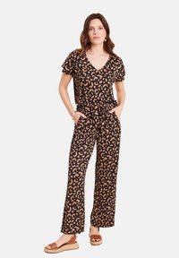 Schwarzer Jumpsuit mit kurzen, rüschigen Ärmeln, einem beigen Tiermuster, V-Ausschnitt, Gürtel in der Taille und weitem Schnitt, kombiniert mit hellbraunen Espadrilles.