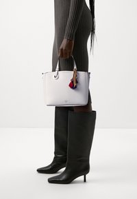 Osoba w prążkowanej sukience trzymająca białą torebkę z kolorowymi frędzlami, nosząca czarne, wysokie na kolana, obcasy na jednolitym tle.