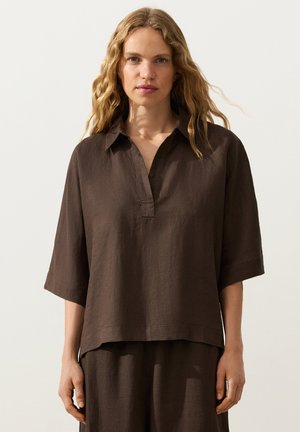 Femme aux longs cheveux blonds ondulés, vêtue d'une chemise ample en lin marron foncé à manches courtes et d'un pantalon assorti, debout devant un fond clair uni.