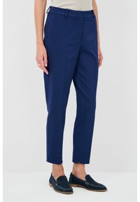 Marineblauwe op maat gemaakte broeken met een slim fit, voorzien van zijzakken en kleine splitjes bij de enkel. Gedragen met donkerblauwe suède instappers.