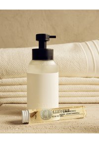 Witte pompflacon met zwarte pompdeksel, naast een heldere glazen fles lichaamswas, geplaatst op opgevouwde crème-kleurige handdoeken. Getextureerde achtergrond.