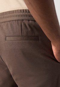 Brune bomullsbukser med elastisk midje, med en baklomme og en jevn tekstur, nærbilde av baksiden.