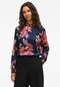 Blusa floreale in satin blu navy con fiori rosa brillante, arancioni e viola. Presenta un colletto alto e maniche lunghe con polsini arricciati.