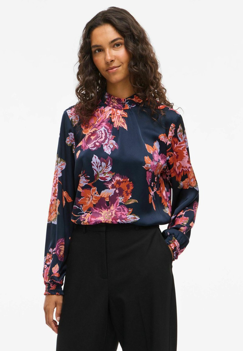 Blusa floreale in satin blu navy con fiori rosa brillante, arancioni e viola. Presenta un colletto alto e maniche lunghe con polsini arricciati.
