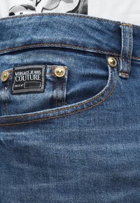 Zili džinsi ar zelta toņu furnitūru, ar melnu etiķeti, kurā rakstīts "VERSACE JEANS COUTURE" un kontrastējošiem oranžiem šuvju detaļām.