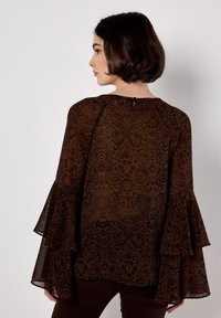 Blusa marrón transparente con intrincados patrones florales negros, cierre en forma de llavero en la parte posterior y mangas acampanadas con volantes en capas.