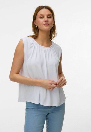 Femme aux cheveux châtain clair portant un chemisier blanc sans manches et un jean bleu clair, debout les mains jointes lâchement.