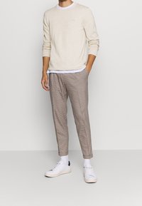 Cremefarbener Strickpullover mit langen Ärmeln, kombiniert mit leichten karierten Hosen in Brauntönen, ergänzt durch weiße Turnschuhe und Knöchelsocken.