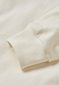 Tessuto color crema con una texture a coste, caratterizzato da un dettaglio con maniche a polsino. Materiale liscio e morbido con sottili linee verticali.