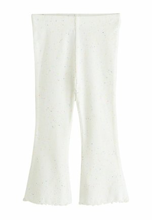 Jeune fille portant un haut rose à manches volantées et un pantalon blanc évasé, chaussée de sandales blanches à boucle, sur un fond beige.