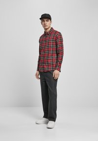 Rødt og svart rutete skjorte med knapper, tettsittende design, kombinert med mørkegrå løse bukser og hvite joggesko. En enkel svart caps fullfører antrekket.