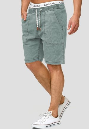 Mann trägt grüne Shorts mit Kordelzug und großen vorderen Taschen, sichtbarem Calvin Klein Bund und weißen Sneakern, steht auf einem Bein.