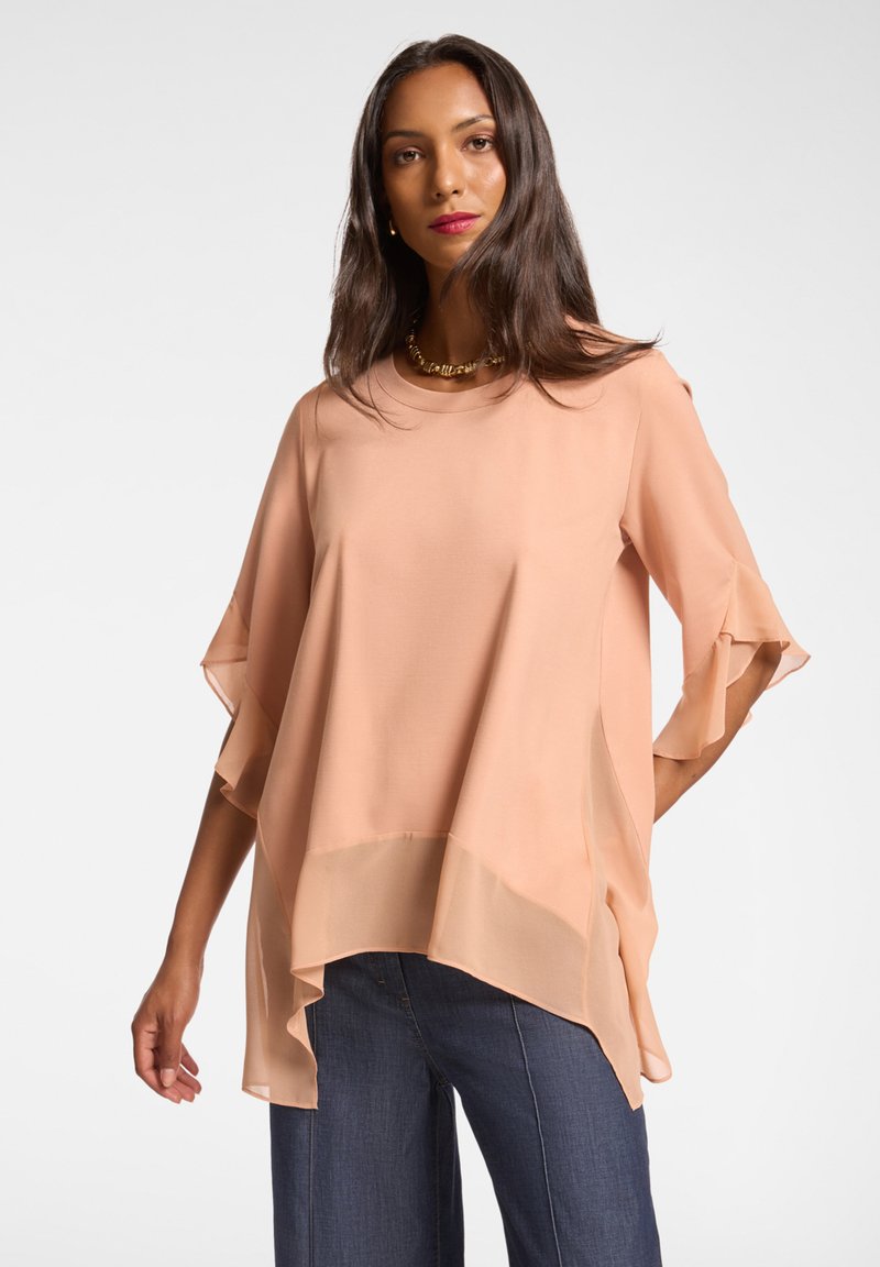 Hedvábná blůza meruňkové barvy, lehká, s vykrojenými rukávy a asymetrickým lemováním. Vyrobena z měkkého materiálu s volným střihem. Kombinována s tmavými džíny.