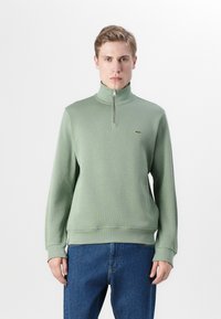 Groene pullover sweatshirt met een halve ritssluiting, geribbelde textuur en een klein geborduurd logo op de borst, gecombineerd met blauwe jeans.
