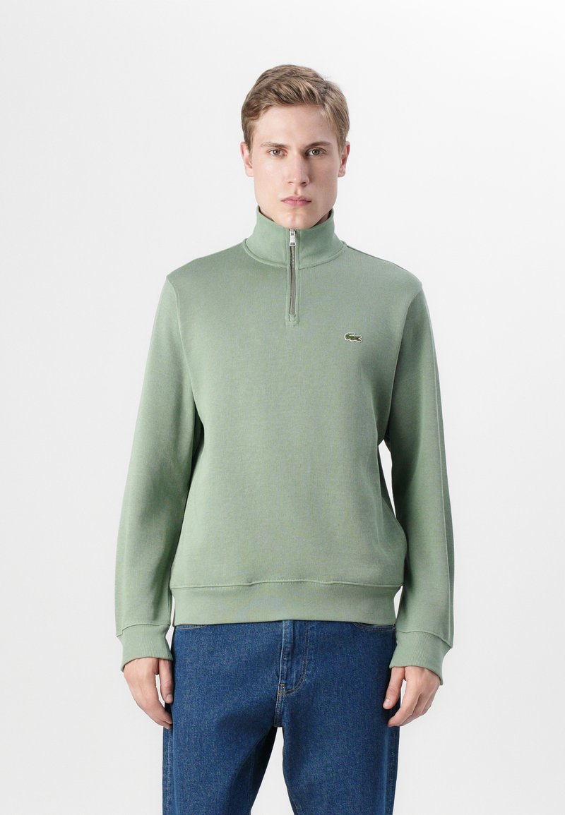 Groene pullover sweatshirt met een halve ritssluiting, geribbelde textuur en een klein geborduurd logo op de borst, gecombineerd met blauwe jeans.