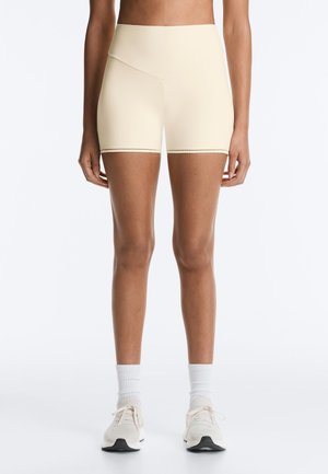 Femme portant un short de sport beige taille haute, des chaussettes blanches montantes et des chaussures de course beiges, debout devant un fond blanc uni.