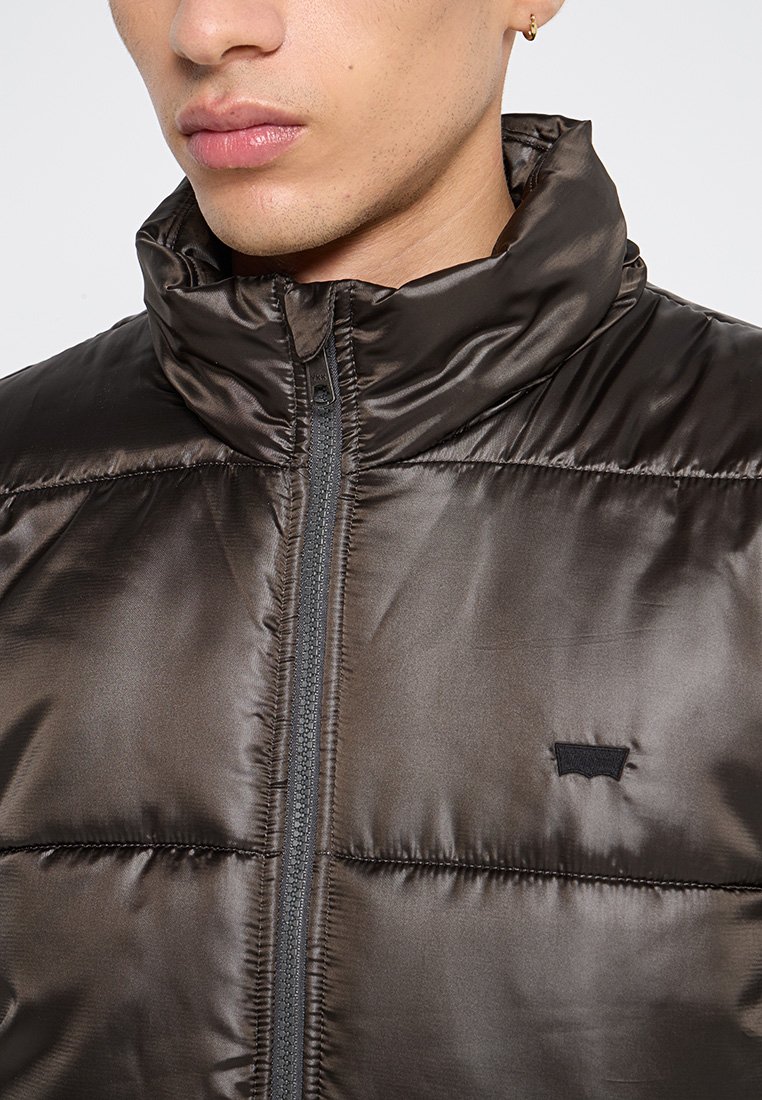 Vista all'altezza del petto di una giacca a vento marrone con una texture liscia e lucida, zip completa, colletto alto e un piccolo logo nero sul lato sinistro.