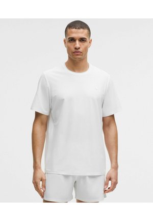 Man met kort haar, gekleed in een eenvoudig wit T-shirt met korte mouwen en witte korte broek, staand tegen een effen lichte achtergrond.