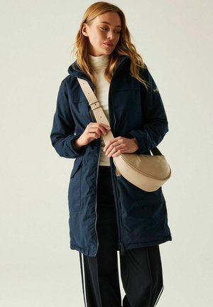 Kobieta w granatowym płaszczu i czarnych spodniach poprawiająca pasek beżowej torby na ramię, stojąca na tle jasnego, jednolitego tła.