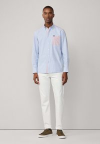 Camicia button-up a righe di colore azzurro chiaro con una tasca a quadretti rosa, abbinata a pantaloni bianchi e sneakers marroni. Tessuto dalla texture liscia.