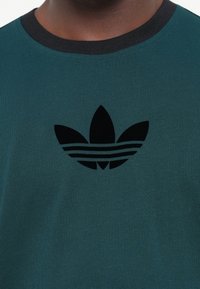 Teal t-shirt med et sort, gummieret Adidas-logo med tre striber. Rund hals med en kontrasterende sort krave, lavet af blødt bomuldsblanding.