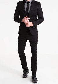 Musta puku räätälöidyllä istuvuudella, jossa on lovellinen kaulus, valkoinen pilkullinen paita ja musta solmio. Kiiltävät mustat juhlakengät täydentävät asun.