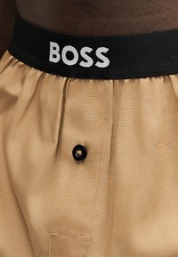 Gros plan sur un boxer couleur beige avec une ceinture noire comportant un texte blanc "BOSS" et un bouton noir sur la braguette avant.