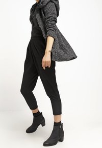 Cappotto grigio strutturato con cappuccio, top nero e pantaloni neri plissettati e affusolati. Stivali neri robusti alla caviglia con un tacco piccolo.