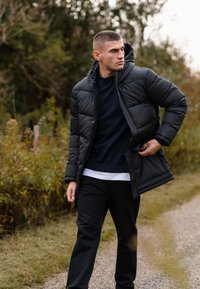 Sort polstret frakke med hætte, med lynlås foran og lommer, båret over en mørk sweater og en hvid T-shirt, parret med sorte bukser.