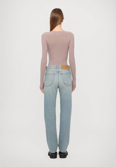 Langärmliges, mauvefarbenes Oberteil kombiniert mit hellblauen, hochgeschnittenen Jeans, die ein braunes Lederpatch und eine lockere Passform haben. Schwarze klobige Schuhe.