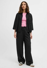 Chemise noire surdimensionnée à boutons sur un débardeur rose côtelé, associée à un pantalon noir ample et des baskets en argent. Texture de tissu lisse.