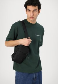 Melns plecu maks ar rāvējslēdzēju nodalījumiem, valkāts uz pleca virs zaļas T-kreklu ar baltu tekstu, kopā ar ziliem džinsu biksēm.