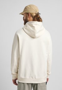 Cremefarbener Hoodie mit lockerer Passform, gerippten Bündchen und einer großen Kapuze. Der Stoff wirkt weich mit einer strukturierten Webart. Kombiniert mit einer hellbeigen Kappe.
