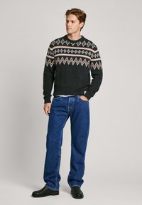 Maglione nero lavorato a maglia con motivo geometrico bianco e rosa, abbinato a jeans di denim blu larghi e stivali neri alla caviglia.