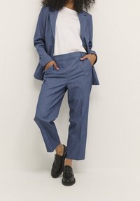 Blå skräddarsydd kavaj med slagkrage, matchad med pleated byxor. Bärs över en vit t-shirt. Svarta loafers fulländar looken.