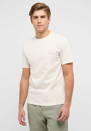 Junger Mann mit hellbraunen Haaren, der ein schlichtes, off-white kurzärmeliges T-Shirt und hellgrüne Hose trägt und vor einem einfarbigen Hintergrund steht.