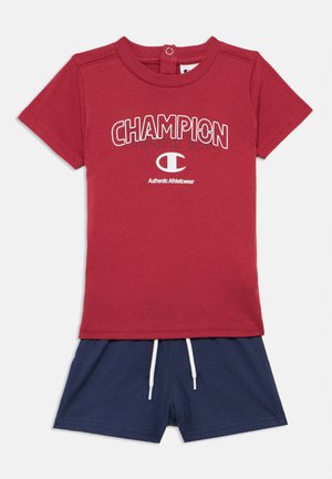 Camiseta roja de manga corta de Champion con logo y texto en blanco, combinada con pantalones cortos azul marino con cordones blancos.