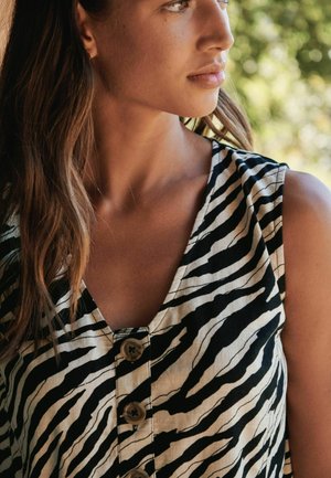 Young woman with long brown hair wearing a sleeveless zebra-striped buttoned top, looking to the right against a blurred outdoor background.