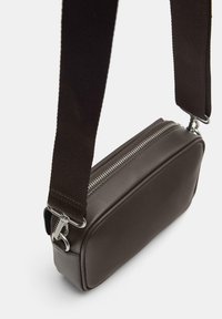 Borsa a tracolla in pelle marrone con forma rettangolare, cerniera argentata e tracolla in tessuto scuro. Texture liscia con elementi hardware minimi.