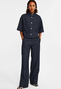 Mörk denim tvådelad outfit bestående av en kortärmad skjorta med knappar och två framfickor samt vida byxor med vertikala ränder.