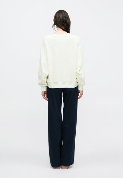 Φορμάκι υπερμεγέθες, ανοιχτό κρεμ, με στρογγυλή λαιμόκοψη και ριγμένα μανίκια. Συνδυάζεται με σκούρο ναυτικό παντελόνι φαρδιάς γραμμής. Λεία υφή.